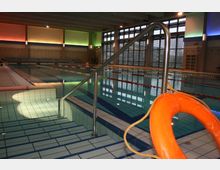 Ein Hallenbad mit einem Schwimmbecken, das durch Leinen in Bahnen unterteilt ist. Im Vordergrund ist ein Handlauf an einer Einstiegstreppe sowie ein Rettungsring zu sehen, im Hintergrund sind große Fenster und farbige Wandbeleuchtung erkennbar.