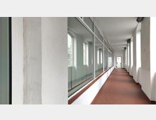 Langer, schmaler Korridor mit einem Boden aus rotbraunen Fliesen, links große Fensterfronten mit Glas und rechts eine Wand mit hohen, schmalen Fenstern. Am Ende des Korridors ist eine Glastür sichtbar.