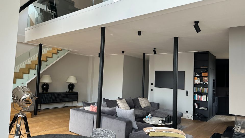 Offener, moderner Wohnbereich mit hoher Decke und Galeriegeschoss mit Glasgeländer, verbunden durch eine Holztreppe. Im Raum stehen ein großes graues Ecksofa, ein runder Esstisch und schwarze Stützsäulen; rechts sind ein Einbau-Regal und eine schwarze Küchenzeile zu sehen.