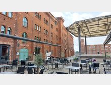 Backsteinbau mit historischen Speichergebäuden, vor denen eine Terrasse mit zahlreichen Sitzgelegenheiten und Sonnenschutzdächern steht. Im Hintergrund weitere Ziegelgebäude und ein freistehender Turm.