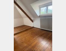 Leerer Dachgeschossraum mit schrägen weißen Wänden, sichtbarem Holzbalken und breiten Holzdielenboden mit kleiner Stufe. Rechts ein Fenster mit Blick ins Grüne über einem großen Rippenheizkörper.