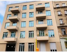 Ein modernes, mehrstöckiges Wohngebäude mit beigefarbener Fassade und symmetrisch angeordneten Balkonen. Die Fenster sind groß und blau getönt, und rechts ist ein angrenzendes, älteres Gebäude mit einer verwitterten Fassade zu sehen.