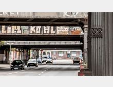 Straßenszene unter einer historischen Eisenbahnbrücke mit verzierten Eisenpfeilern, Autos fahren auf der mehrspurigen Fahrbahn darunter. Die Brücke zeigt Graffiti, und im Hintergrund sind Backsteingebäude zu erkennen.
