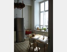 Helle Küche mit schlichtem Design: Ein Esstisch mit vier Stühlen steht in der Mitte, daneben ein großer silberner Kühlschrank. Die Fensterbank vor einem großen Fenster ist mit Pflanzen und Küchenutensilien dekoriert. Der Boden hat ein gemustertes Fliesendesign.