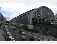 Großer Bahnhof mit moderner Glasfassade und halbrundem Dach, flankiert von breiten Gehwegen und mehrstöckigen Gebäuden. Vor dem Eingang befinden sich geparkte Autos, Fahrräder und Fußgänger.