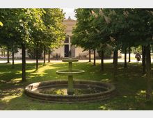 Ein kleiner Park mit einer symmetrischen Baumreihe, in deren Mitte ein grüner, runder Brunnen mit einer Statue steht. Im Hintergrund ist ein historisches Gebäude mit heller Fassade und einem großen Eingang zu sehen.