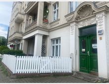 Alt-Text: Historisches Wohngebäude mit verzierter weißer Fassade und grüner Doppeltür als Eingang. Vorgarten mit weißem Lattenzaun, Balkone mit schmiedeeisernen Details, und gepflasterter Gehweg im Vordergrund.