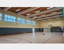 Innenansicht einer großen Sporthalle mit markiertem Hallenboden, hohen Fensterreihen entlang der Seitenwand und einer Holzdecke mit langen Deckenleuchten. An der Stirnseite sind ein Basketballkorb und ein Handballtor vor gepolsterten Wandflächen sowie Sitzbänke entlang der Wand zu sehen.