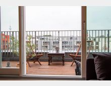 Blick durch eine offene Glastür auf einen Balkon mit Holzboden, zwei Holzstühlen, einem kleinen Tisch mit einer Buddha-Kopf-Skulptur und einer Topfpflanze. Im Hintergrund sind moderne Wohnhäuser und der Berliner Fernsehturm sichtbar.