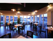 Ein modernes Café mit Holzboden, dunklen Stühlen und Tischen, die mit Tischdecken dekoriert sind. Große Fensterfronten bieten Ausblick auf eine Außenanlage mit Brücke, Wasser und Reihenhäusern.