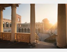 Blick von einem Säulengang auf einen historischen Innenhof mit Arkaden, Steinbalustraden und einer Treppenanlage, beleuchtet von tiefstehender Sonne im Hintergrund. Im Hof sind weitere Säulen und eine Skulptur erkennbar, umgeben von Bäumen.