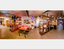 Innenraum eines Ausstellungs- oder Lagerraums in einem Kellergewölbe mit weiß gestrichenen Ziegelbogen-Decken und Betonboden. In der Mitte stehen lange Tische und Metallregale mit alten technischen Geräten, Leuchten und Schildern; rechts führt ein Durchgang in einen weiteren Raum.