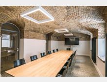 Moderner Konferenzraum in einem historischen Gewölbe mit Backsteinmauerwerk. Der Raum verfügt über einen langen Holztisch, schwarze Stühle, rechteckige Hängelampen, einen großen Bildschirm und gewölbte Fenster sowie Türen.