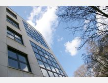 Moderne Fassade eines mehrstöckigen Gebäudes mit großen, rechteckigen Fenstern, aufgenommen aus einem niedrigen Winkel. Im Hintergrund sind ein blauer Himmel mit weißen Wolken und einige kahle Äste von Bäumen zu sehen.