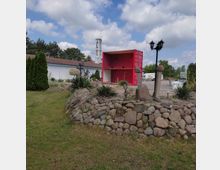 Eine Grünfläche mit einem Steingarten im Vordergrund, dekoriert mit Pflanzen und Steinskulpturen. Im Hintergrund stehen ein roter ausrangierter Schiffscontainer, eine weiße Gebäudeanlage mit Flachdach und ein hoher Industrieschornstein; der Himmel ist teils bewölkt.