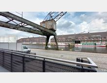 Historischer Hafengebäudekomplex mit Backsteinfassaden an einem Wasserkanal, im Vordergrund ein alter Hafenkran aus Metall und gepflasterte Hafenpromenade, im Hintergrund liegen weiße Tanks und ein Ausflugsschiff am Ufer.