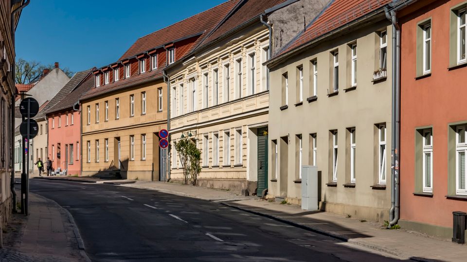 Curving residential street lined with closely spaced, two- to three-story pastel stucco row houses with red-tiled roofs and white-framed windows. Narrow sidewalks, street signs, and utility pipes run along the building facades under a clear blue sky.