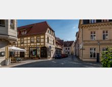 Eine gepflasterte historische Straßenkreuzung mit Fachwerkhäusern und Gebäuden im klassischen Stil. Links vorne steht ein Café mit Tischen und Sonnenschirmen, während geparkte Autos die Straße säumen.