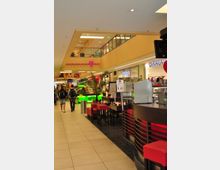 Eine Innenansicht eines Einkaufszentrums mit Geschäften auf zwei Etagen. Im Vordergrund befinden sich Tische und Stühle eines Cafés mit roten Polstern, daneben grüne Leuchtschilder und Pflanzen.