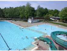 Freibad mit einem großen Schwimmbecken, einem flachen Kinderbecken und einer grünen Wasserrutsche im Vordergrund. Im Hintergrund sind Bäume, ein Umkleidegebäude und Liegebereiche mit Personen unter Sonnenschirmen zu sehen.