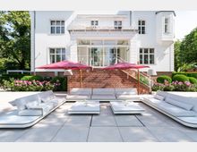 Weiße Villa mit Backsteinsockel und einer Terrasse, die durch eine Treppe mit filigranem Metallgeländer zugänglich ist. Im Vordergrund moderne, helle Sitzmöbel und niedrige Tische auf einer gepflasterten Fläche, ergänzt durch rote Sonnenschirme und umliegende, gepflegte Büsche und Blumenbeete.