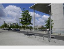 Offene städtische Szenerie mit breitem gepflastertem Gehweg, einzelnen Bäumen und moderner Architektur im Hintergrund. Rechts im Bild ist eine große Betonwand mit einem überhängenden Dach zu sehen, während links Autos entlang einer Straße parken.