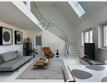 Modernes Wohnzimmer mit weißen Wänden, einem grauen Sofa und einem Ledersessel. Im Raum befinden sich eine Holz-Couchtisch, ein Teppich mit strukturierter Oberfläche, eine Treppe mit Glasgeländer und mehrere große Fenster, die viel Tageslicht hereinlassen.