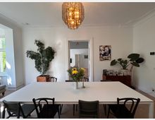 Esszimmer mit weißem Tisch und schwarzen Stühlen, dekoriert mit einer Vase mit Blumen in der Mitte. Im Hintergrund eine weiße Wand mit Gemälde, Zimmerpflanzen und einer Kommode mit gerahmten Fotos, ergänzt durch großen Kronleuchter an der Decke.