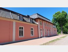 Ein langgestrecktes, rosa Gebäude mit weißen Fensterrahmen und einem dunklen Schieferdach, das mehrere Gauben enthält. Im Vordergrund befindet sich ein gepflasterter Weg, und im Hintergrund sind grüne Bäume zu sehen.