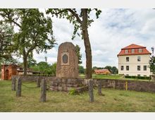 Ein steinernes Denkmal mit einer Inschriftplatte steht auf einem Sockel, umgeben von einer Kette und Pfosten, in einer Grünfläche mit Bäumen. Im Hintergrund sind ein gelbes Gebäude mit rotem Dach und weitere landwirtschaftliche oder historische Gebäude sichtbar.