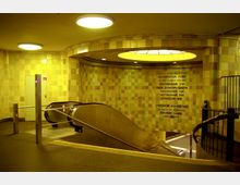 Untergrundbahnhof-Eingang mit gelb-beige gefliesten Wänden, einer großen Leuchte in der Decke und einer Rolltreppe, die zu den Bahnsteigen führt. An der Wand neben der Treppe befindet sich ein Orientierungstext mit Richtungsangaben zu verschiedenen Bahnstationen.
