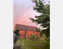Roter Backsteinbau mit mehreren Holztüren und kleinen Fenstern, umgeben von einer Wiese und Bäumen; darüber ein Regenbogen am rosa gefärbten Himmel bei Sonnenuntergang. Im Vordergrund Gartenmöbel und Pflanzen.