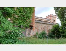 Verfallenes historisches Gebäude mit Backstein- und Putzfassade, teils überwuchert von Bäumen und Pflanzen. Das Gebäude zeigt dekorative architektonische Elemente wie Gesimse und einen kleinen Turm mit Zinnen.