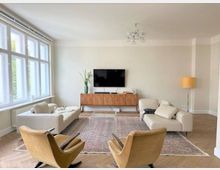 Helles Wohnzimmer mit großen Fenstern auf der linken Seite, zwei cremefarbenen Sofas und zwei gelben Sesseln, einem Teppich auf Holzfußboden sowie einer Wand mit Flachbildfernseher und einer Holzkommode. Eine Stehlampe mit gelbem Schirm steht rechts neben der Kommode.