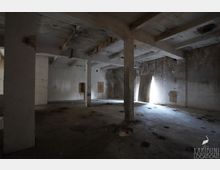 Großer, leerer Betonraum in einem verlassenen Industriegebäude mit massiven Stützpfeilern, niedriger Decke und fleckigen, abgenutzten Wänden. Seitlich fällt helles Tageslicht durch eine schmale Öffnung ein und wirft Schatten auf den staubigen Boden mit mehreren runden Vertiefungen.