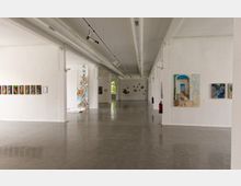 Großer, moderner Ausstellungsraum mit weißen Wänden, poliertem Betonboden und zahlreichen Kunstwerken an den Wänden und auf Podesten. Die Decke ist mit sichtbaren Lüftungsrohren und Schienensystemen für Beleuchtung ausgestattet.