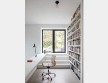 Minimalistisches Arbeitszimmer mit weiß gestrichenen Wänden, einem Einbauschreibtisch, einem Holz-Drehstuhl und einer großen, deckenhohen Bücherwand rechts. Ein großes Fenster mit schwarzem Rahmen bietet Blick auf einen Garten mit Bäumen.