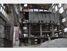 Innenansicht eines verlassenen Fabrikgebäudes mit hohen Betonpfeilern, offenen Stahltreppen und großen, teilweise zerstörten Fenstern. Der Raum ist stark gealtert, mit bröckelnden Wänden, freiliegenden Stahlstrukturen und Schutthaufen am Boden.