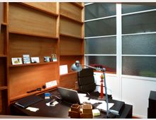 Ein modernes Büro mit einem Schreibtisch aus dunklem Holz, darauf ein Laptop, eine Tischlampe und Bürozubehör. Im Hintergrund befinden sich hohe, leere Holzregale und eine Wand mit strukturiertem Glasfensterrahmen.