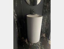 Modernes Badezimmer mit einem freistehenden, weißen, zylindrischen Waschbecken. Dunkle Wände mit Marmoroptik sowie ein runder Spiegel über dem Waschbecken; zwei kleine Tierfiguren aus Metall sind am Boden platziert.