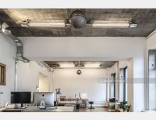 Modernes Büro mit minimalistischer Einrichtung, weißen Wänden und einem Sichtbeton-Deckenstil. Arbeitsplätze mit Monitoren und Schreibtischlampen stehen verteilt, während große Fenster die Umgebung mit natürlichem Licht erhellen.