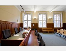 Innenansicht eines Gerichtssaals mit hohen Fenstern, einer hölzernen Wandvertäfelung und einem langen Richtertisch mit schwarzen Bürostühlen. Im Hintergrund befinden sich Wartestühle und eine Holzbank vor einer cremefarbenen Wand.