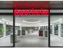 Moderne Apotheke mit großer Glasfront und rotem Leuchtschriftzug „Apotheke“ darüber. Im Innenraum sind helle Regale mit verschiedenen Produkten wie Medikamenten, Pflegeartikeln und Kosmetika gleichmäßig angeordnet.