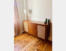 Heller Innenraum mit weißer Wand und breiten Holzdielen; davor steht eine niedrige Holzkommode mit zwei Rattantüren und drei Schubladen. Auf der Kommode befinden sich eine Dreibein-Tischlampe und zwei kleine Kaktus-Dekorationen, links hängt ein bodenlanger Vorhang.