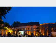 Nachtaufnahme eines historischen Gebäudes aus Stein mit erleuchteten Fassaden, vor dem eine Freiluftveranstaltung mit Publikum und musikalischer Darbietung stattfindet. Auf einer erhöhten Terrasse stehen Menschen mit Fackeln, daneben ein Banner mit der Aufschrift „Glück auf“ und ein Bergbau-Emblem.