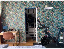 Zimmer mit bunter Wandtapete mit Tropenmuster in Blau- und Grüntönen, einem Holztisch mit Kosmetikartikeln und Spiegel auf der linken Seite sowie einem schwarzen Heimtrainer rechts. Im Hintergrund ist ein offener Kleiderschrank erkennbar.