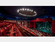 Innenraum eines Theaters mit einer Bühne und gestaffelten Sitzreihen mit roten Polstern. Die Decke ist mit einem sternenhimmelartigen Muster gestaltet, und über der Bühne befindet sich ein rotes Schild mit der Aufschrift "Wintergarten".