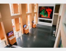 Heller Innenraum eines modernen Gebäudes mit hohen, holzverkleideten Säulen, schwarzen Steinfliesen und großen digitalen Bildschirmen an den Wänden. Ein großer Bildschirm an der Stirnwand zeigt ein farbenfrohes Kunstwerk, während kleinere Bildschirme entlang der Säulen angebracht sind.