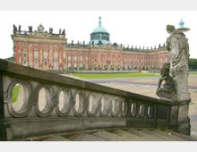 Barockschloss mit roter Fassade, zahlreichen großen Fenstern und verzierten Skulpturen auf dem Dach, im Vordergrund eine steinerne Balustrade mit ovalen Öffnungen und dekorativen Statuen. Die Umgebung umfasst einen offenen, gepflasterten Platz und einen parkähnlichen Bereich.