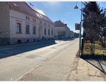 Straße in einer ruhigen Wohngegend mit gepflastertem Gehweg. Links stehen ältere, verputzte Häuser mit roten Ziegeldächern, rechts ein Zaun mit Baumbewuchs.